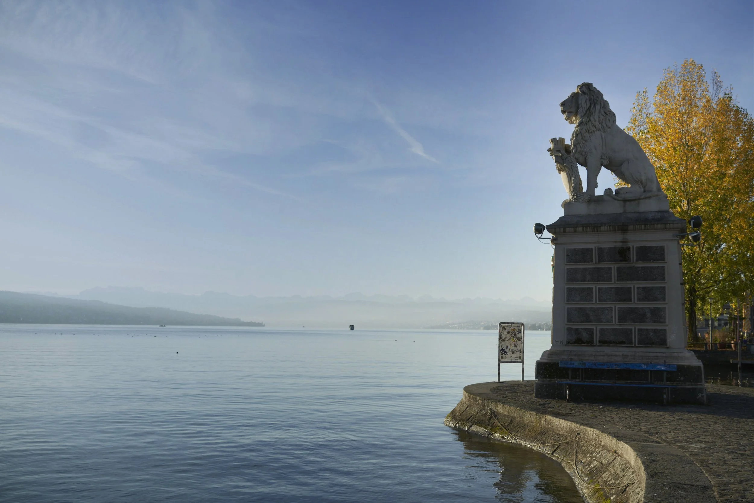 Stone lion statue on a waterfront promenade with calm water and distant mountains under a clear sky.