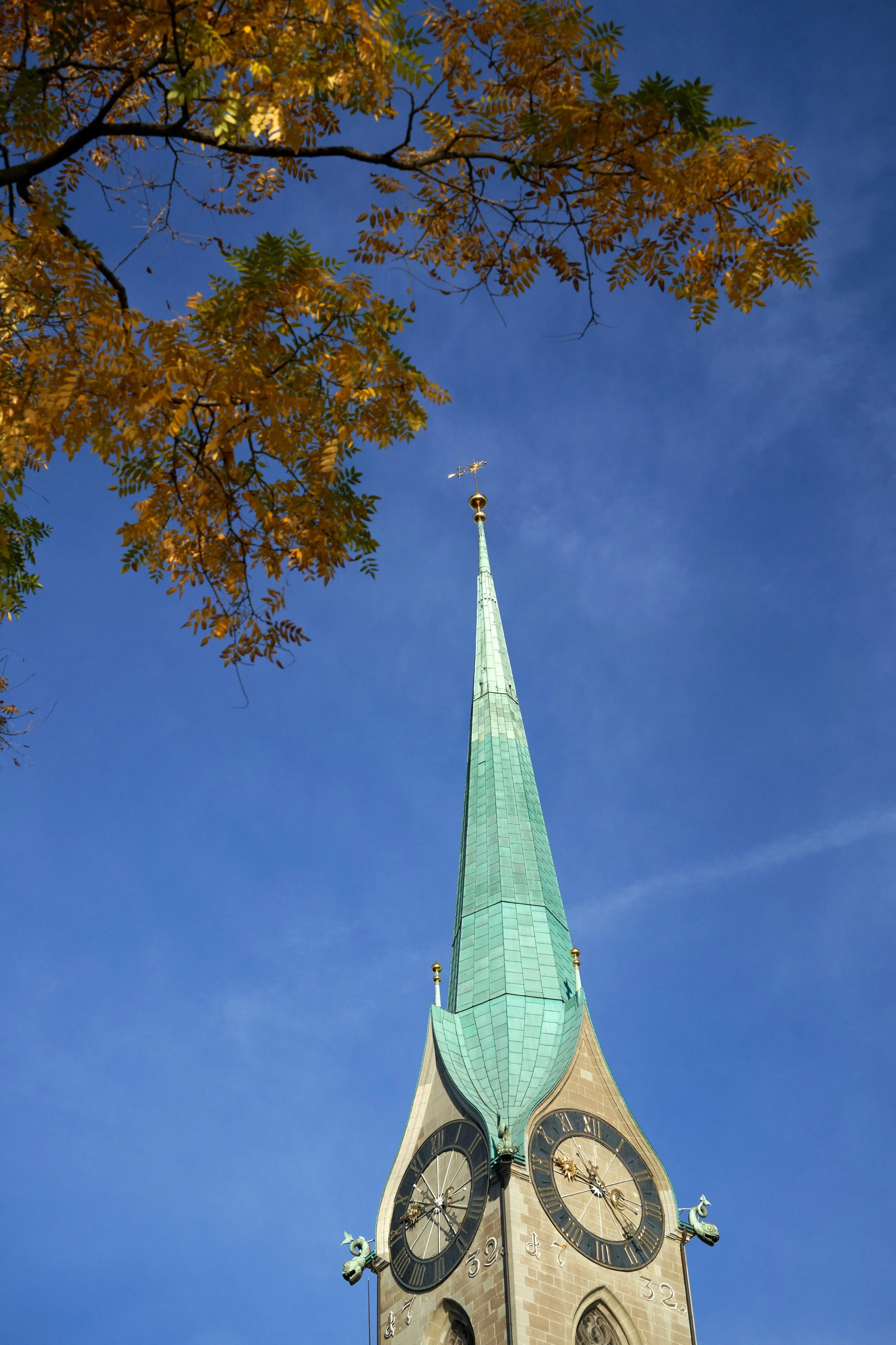 Green spire of a historic clock tower with Roman numeral clock faces under autumn leaves against a clear blue sky.