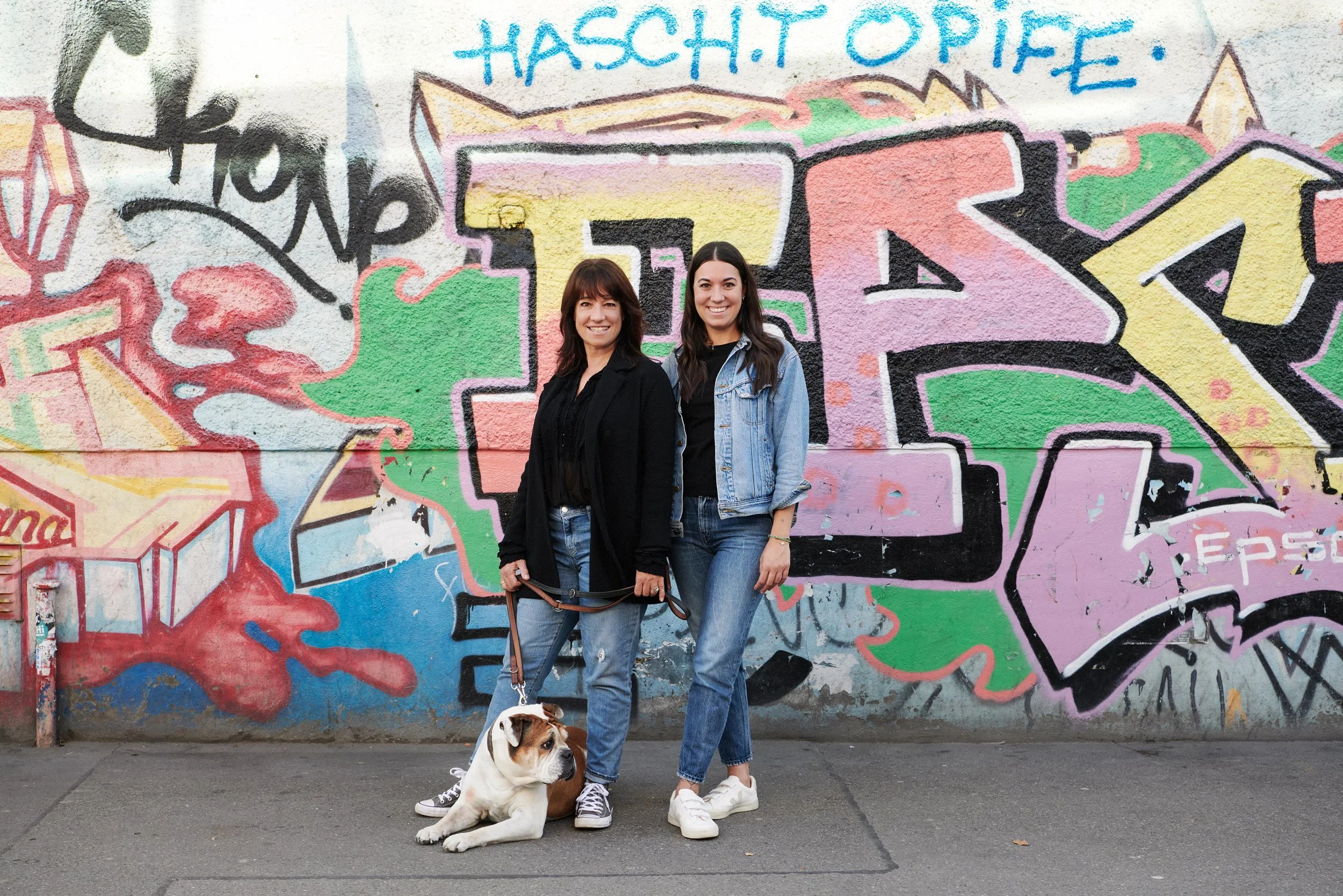 Two women standing in front of a colorful graffiti wall, one holding a leash attached to a sitting bulldog.