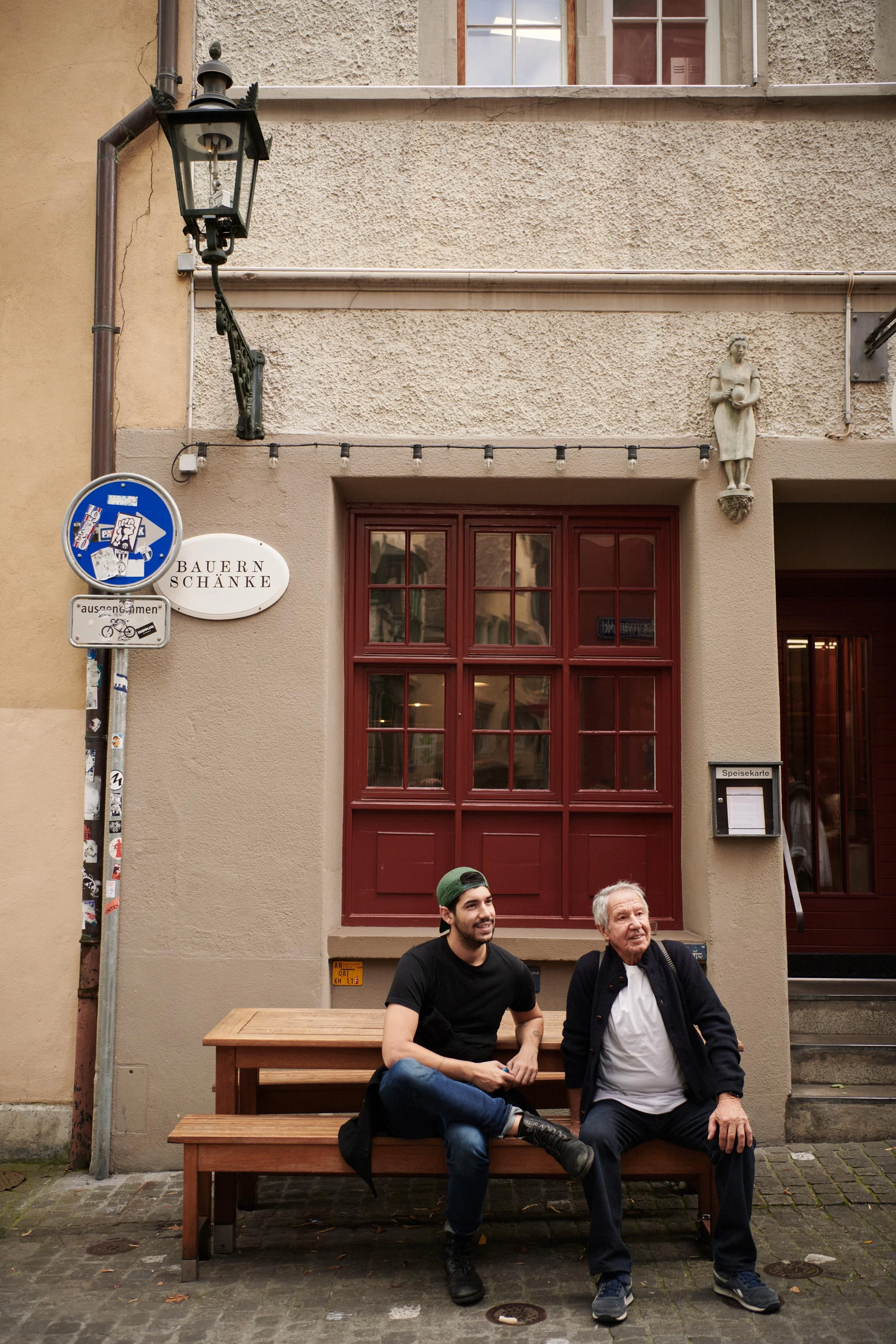 Two men sitting on a wooden bench outside a building with beige walls and red-framed windows.