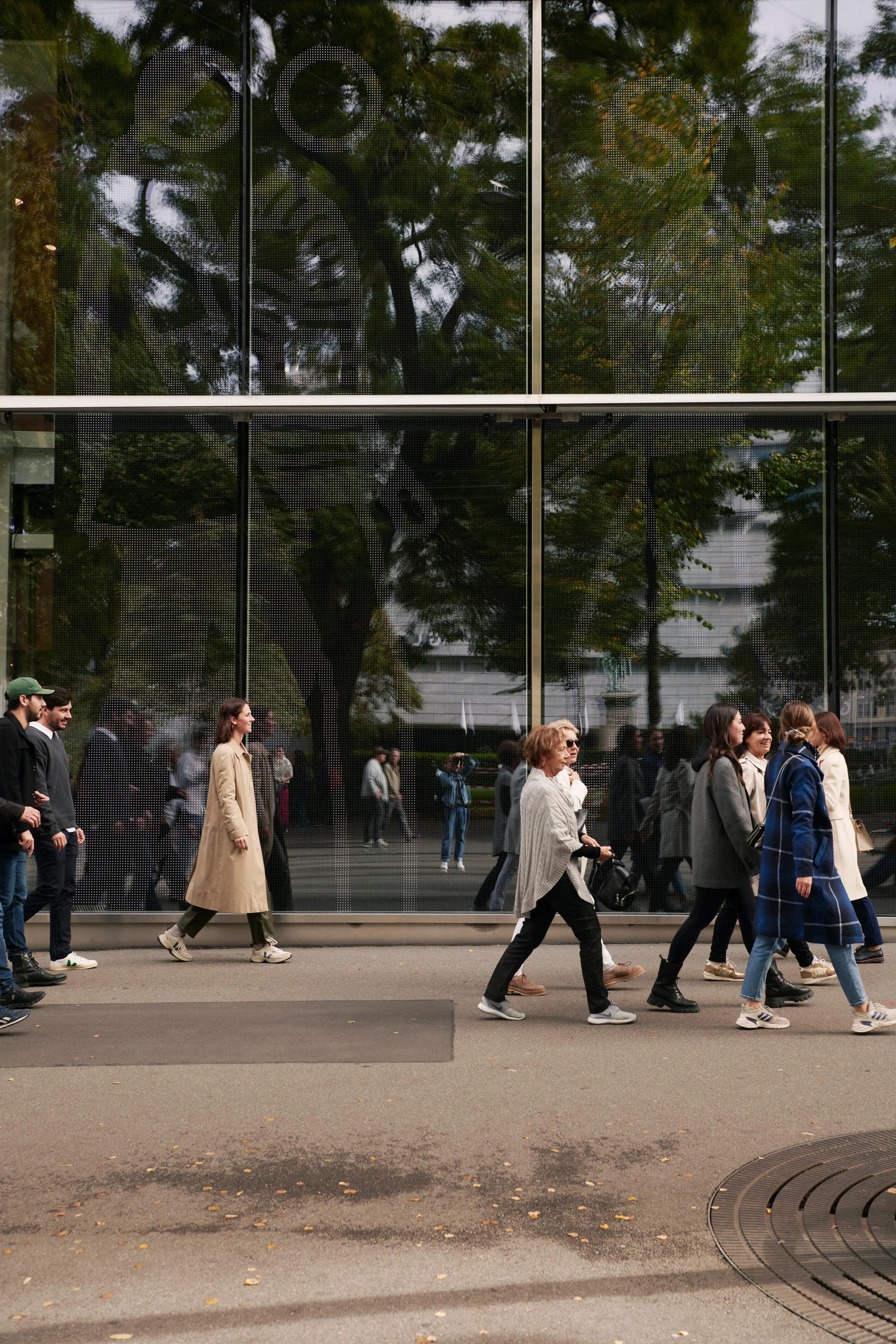 People walking on a sidewalk in front of a large glass wall reflecting trees and urban buildings.