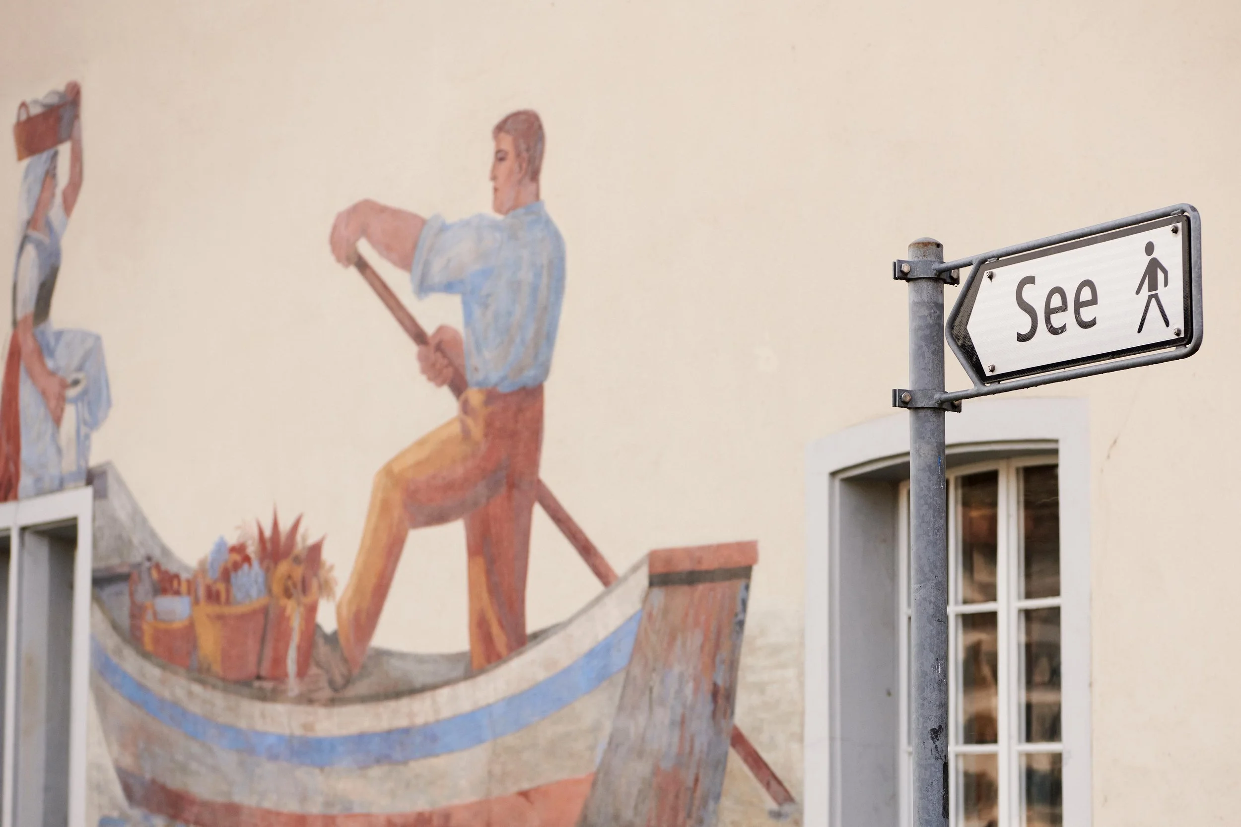 Street sign pointing to See with a walking person icon in front of a wall mural depicting a man rowing a boat with flower baskets and a woman carrying a basket.