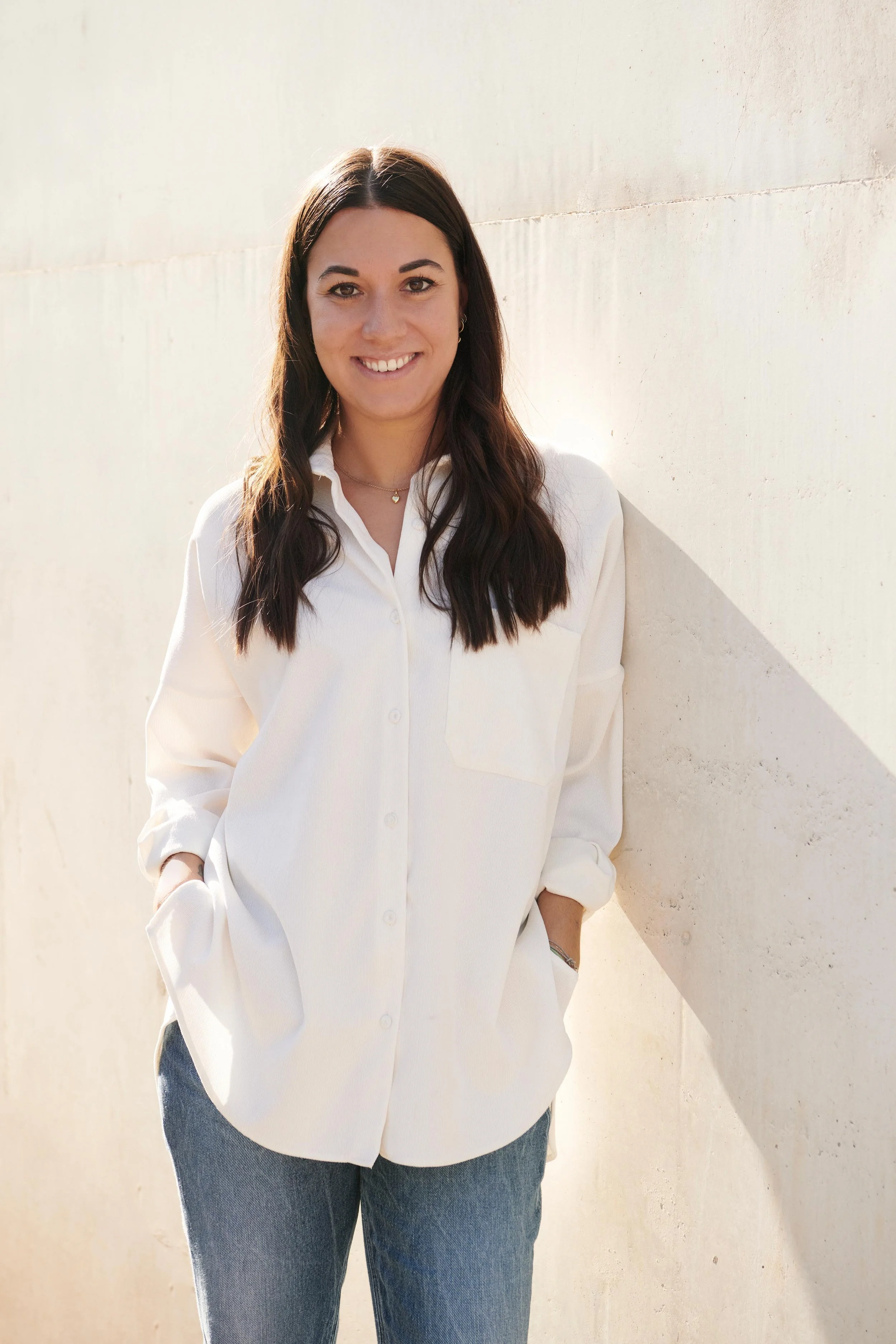 Talisa Arpino - Smiling woman with long brown hair wearing a white button-up shirt and blue jeans, standing against a light-colored wall with hands in pockets.