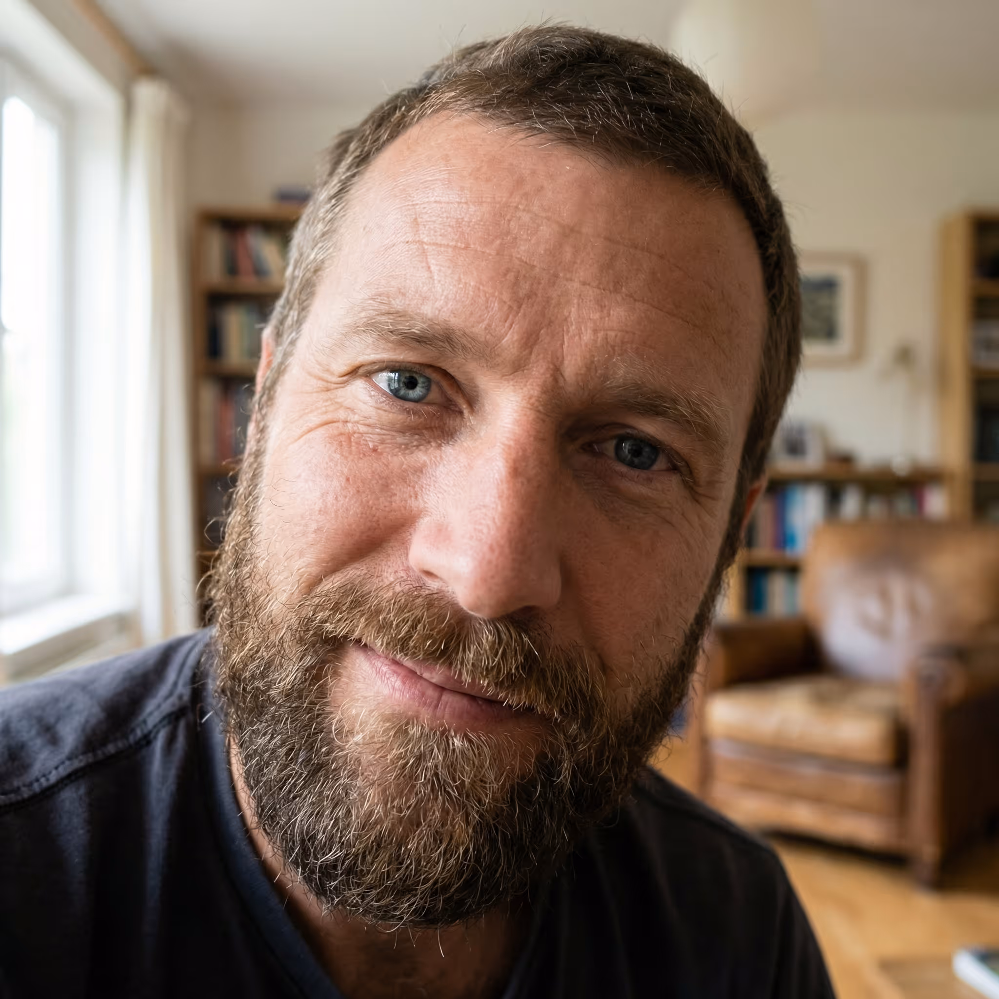 Close-up of a smiling bearded man with blue eyes in a cozy room with a leather armchair and bookshelves.
