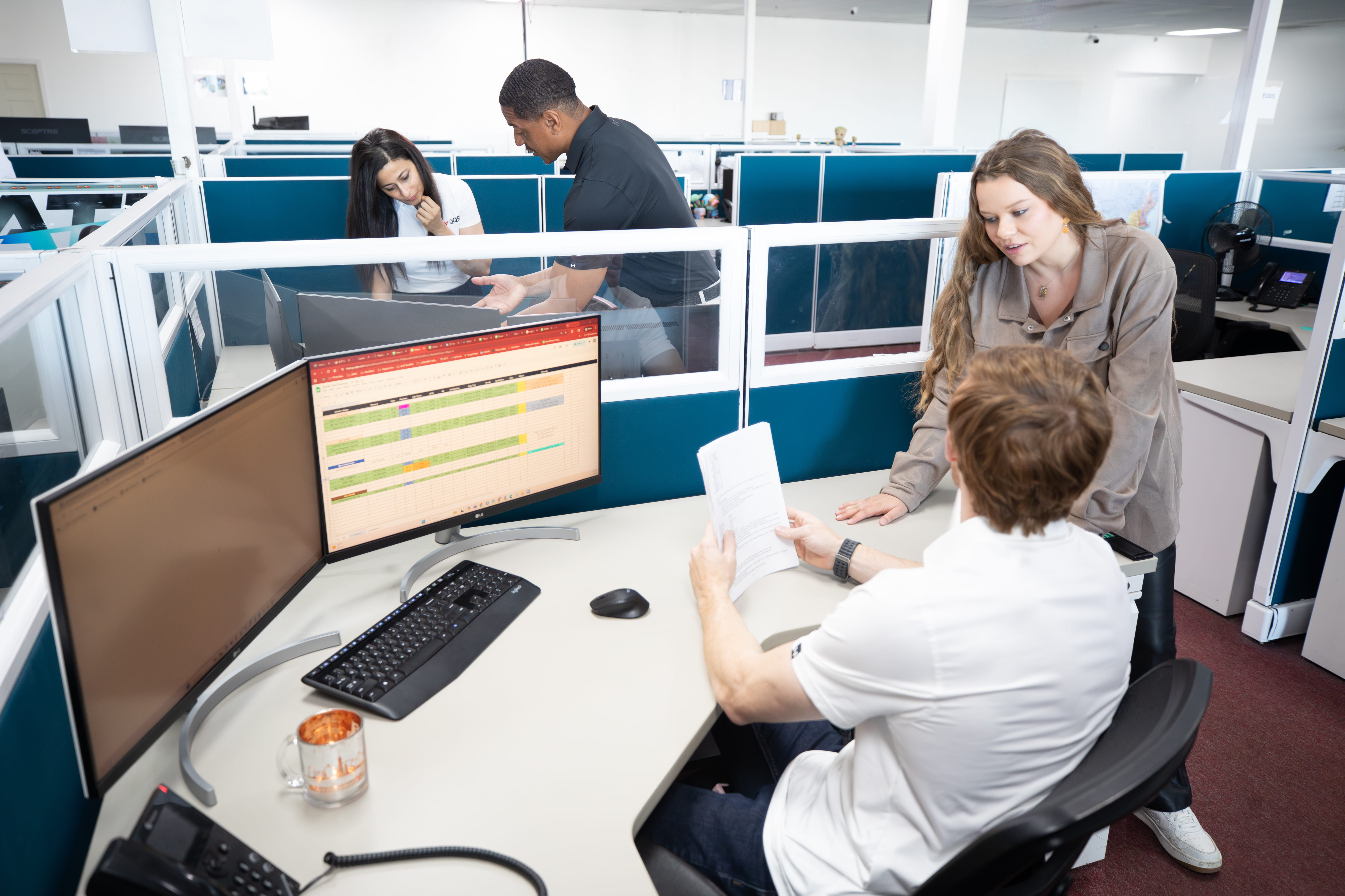 Office workers collaborate at cubicles with computer displays showing data charts and timelines.