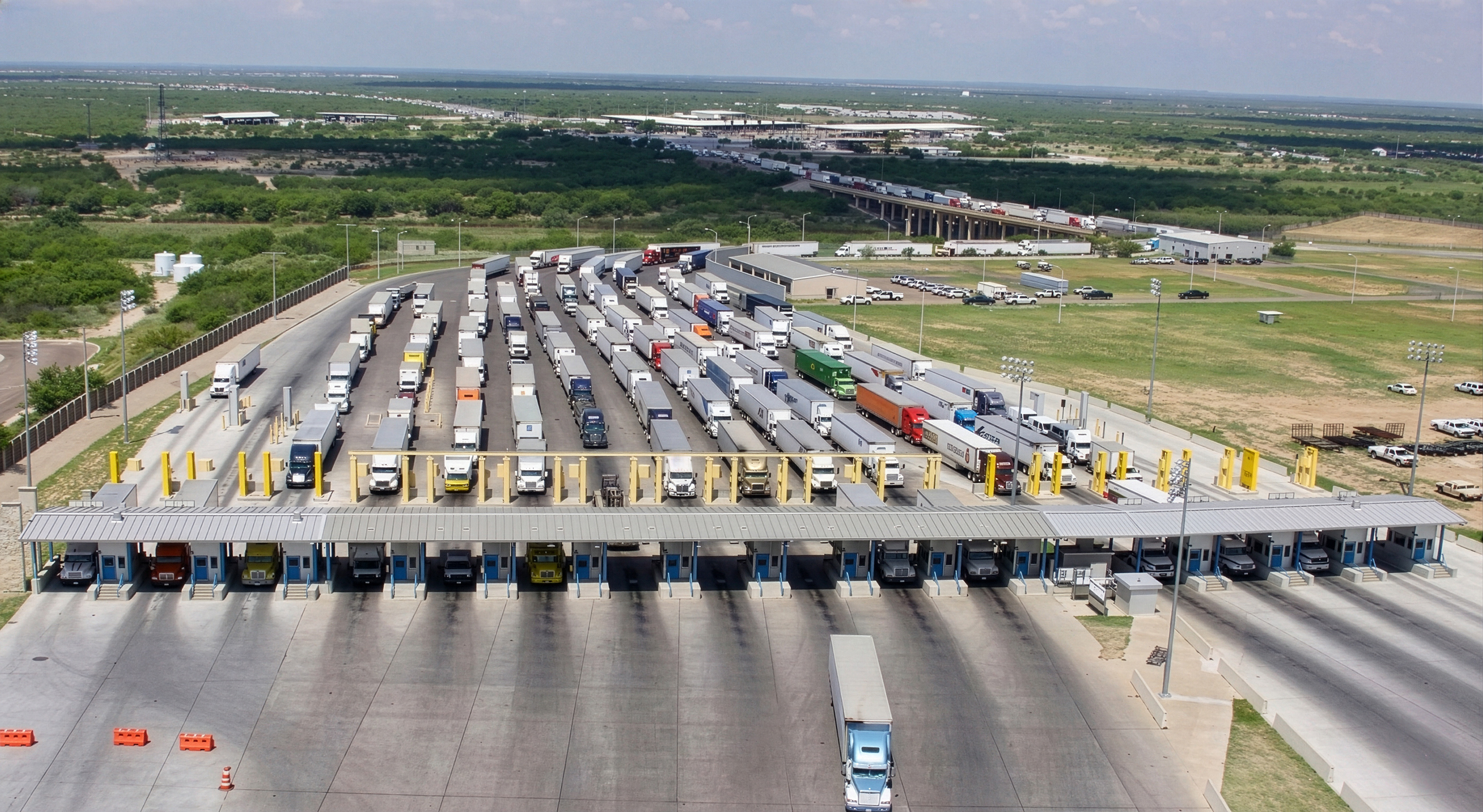 Aerial view of truck inspection facility with multiple lanes and toll booths, parked vehicles and highway.