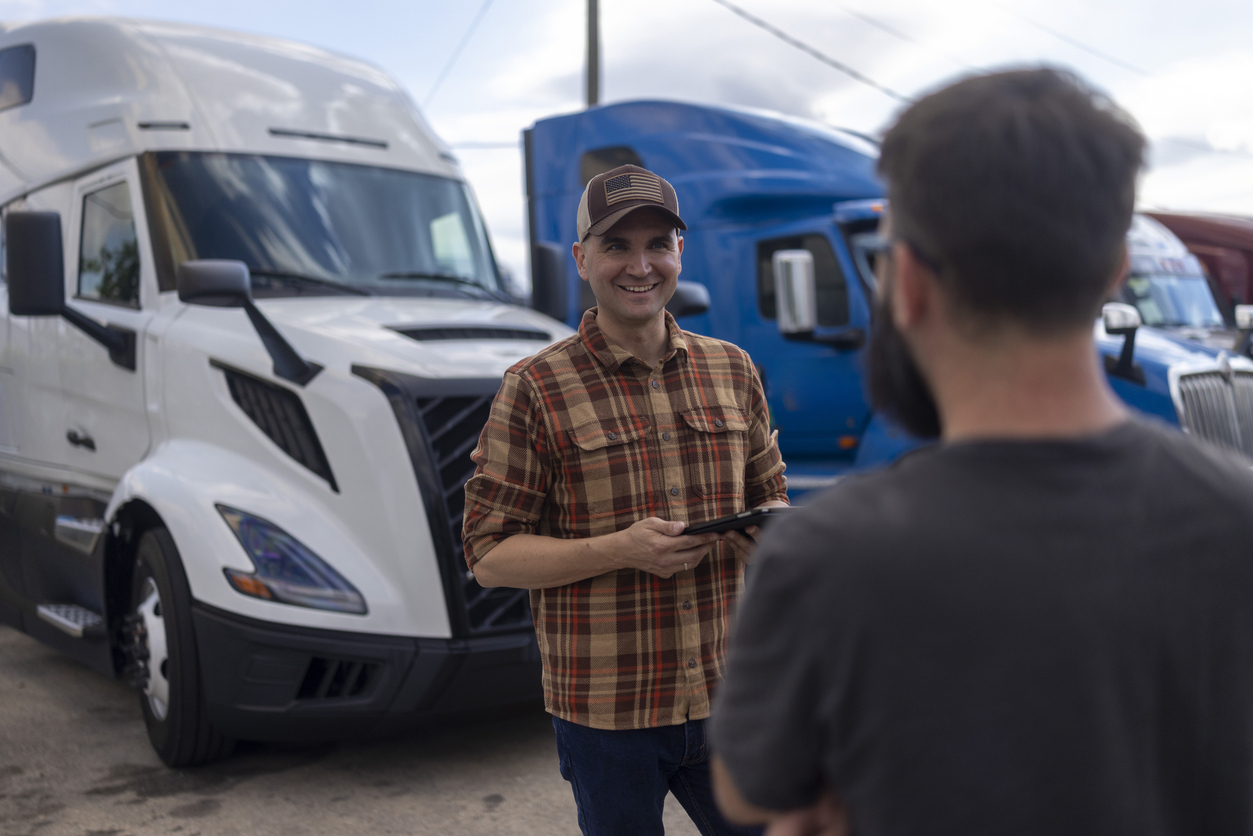 Truck driver in plaid shirt and cap smiling while talking to colleague near parked semi-trucks.