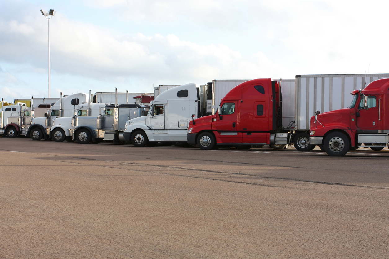 Row of commercial semi-trucks parked in lot, featuring white and red tractors with trailers.