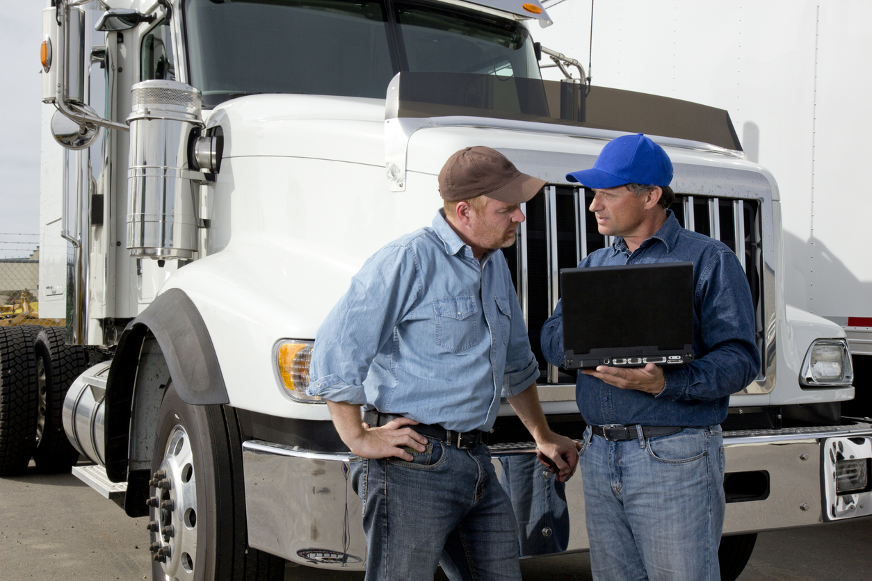 Two truck drivers discuss logistics using laptop next to white semi-truck