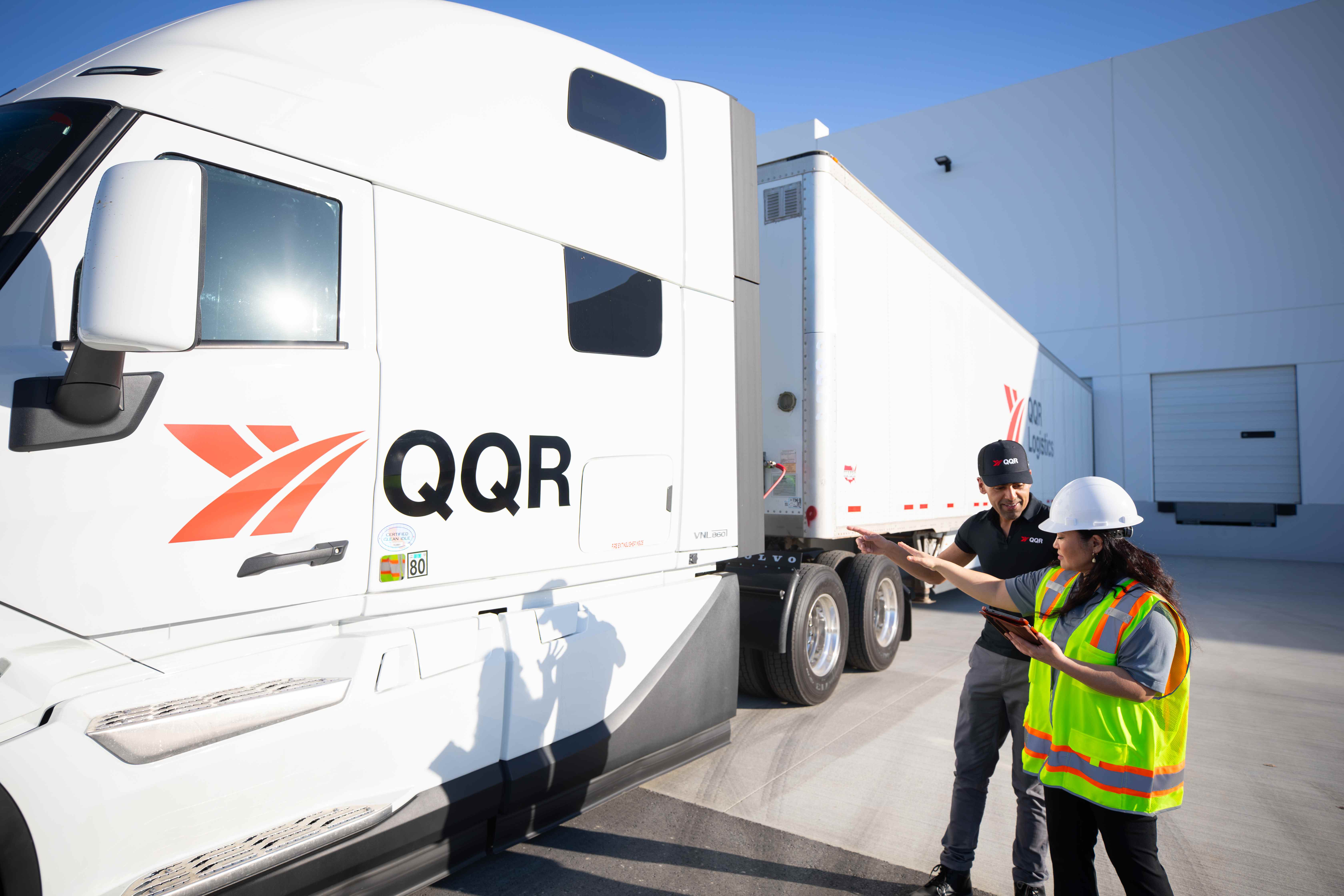 Two workers in safety vests inspecting a white QQR refrigerated transport truck under blue sky.