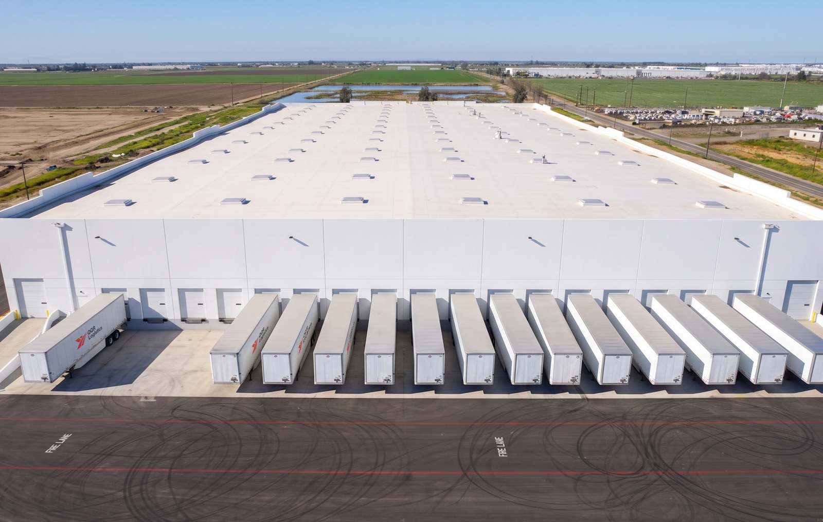 Aerial view of large white warehouse with loading docks and trucks parked in agricultural area