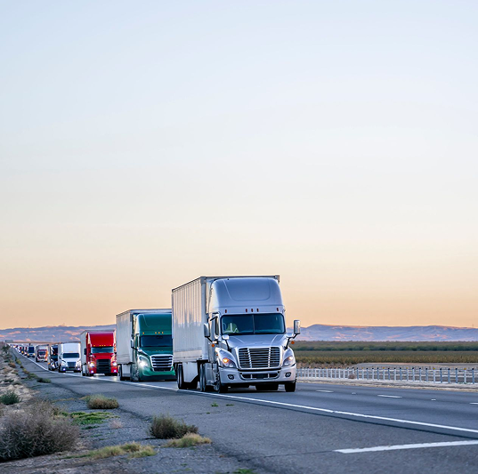 Line of semi trucks driving on highway through desert landscape at sunset.