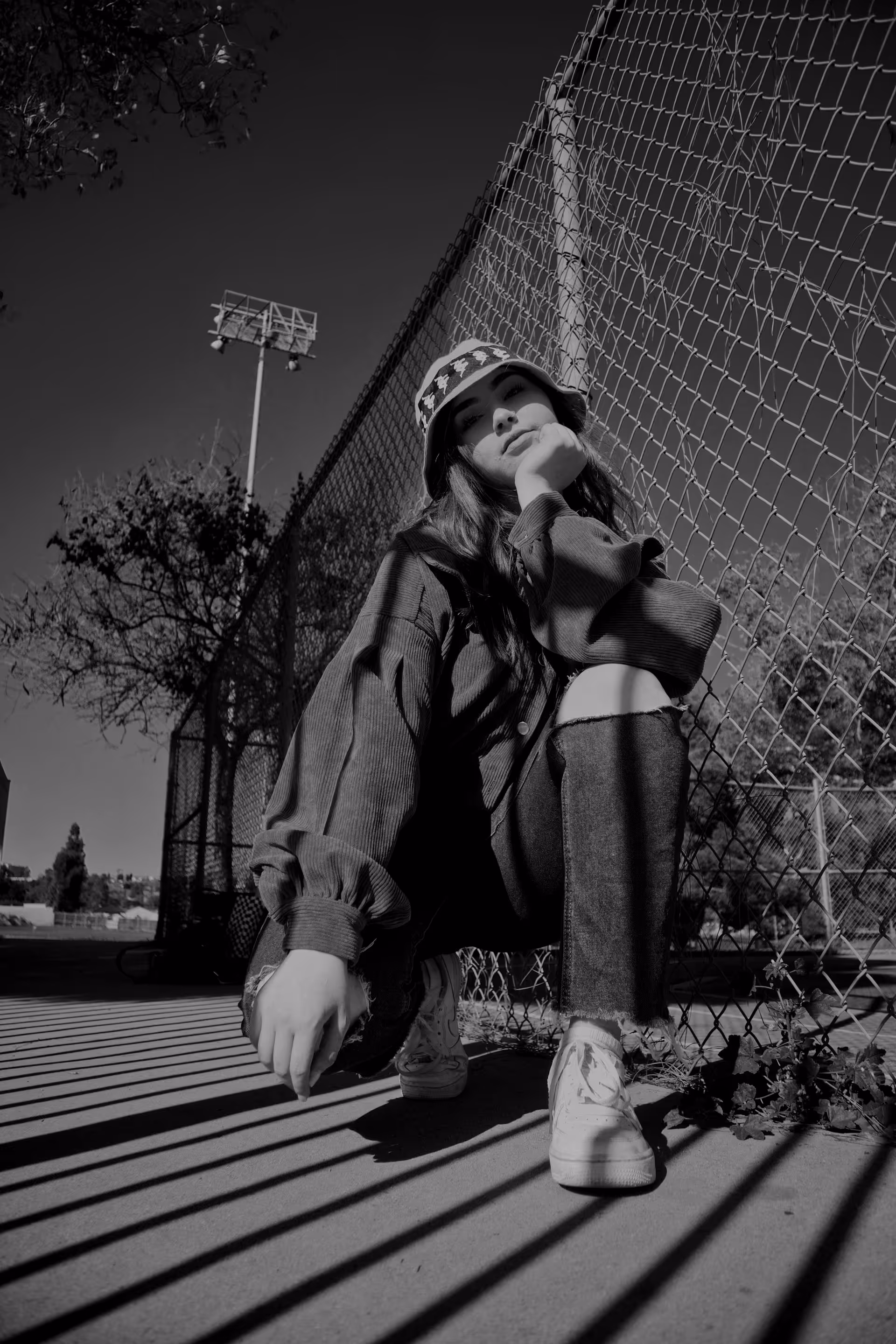 Woman wearing a bucket hat, squatting beside a chain-link fence with shadows cast on the ground.