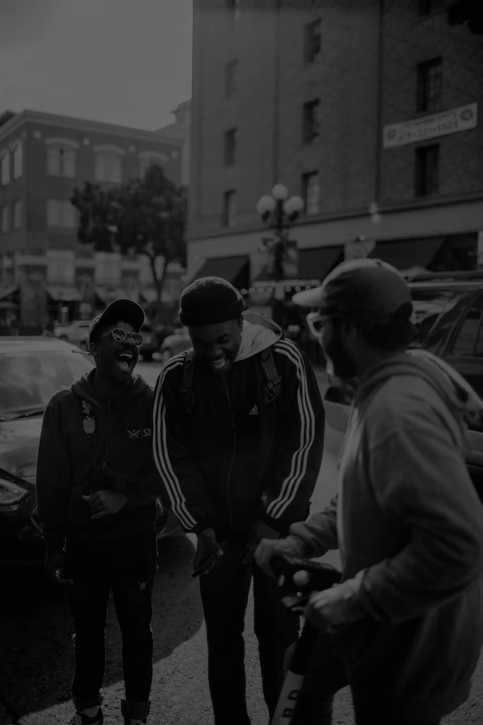 Three men outdoors on a city street, two laughing and one on a scooter.