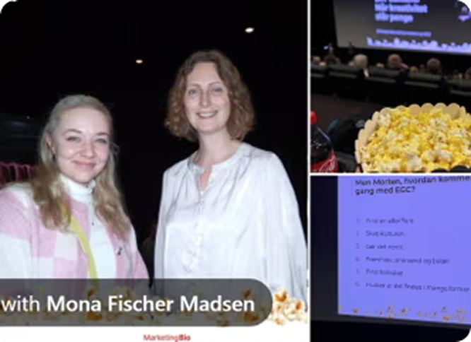 Two smiling women posing indoors at an event, one wearing a pink and white checkered jacket and the other in a white blouse, with a popcorn bucket and a presentation screen visible nearby.