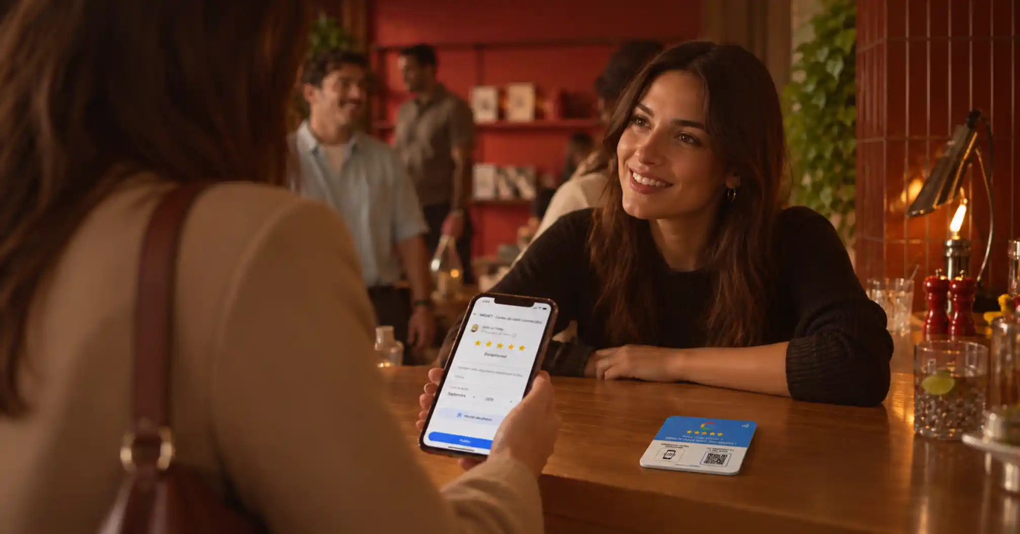 Une femme assise au comptoir d'un café sourit en écoutant une autre femme qui tient un smartphone affichant une page d'avis avec une note cinq étoiles. Sur le comptoir se trouve une plaque NFC bleue avec un QR code pour laisser un avis.