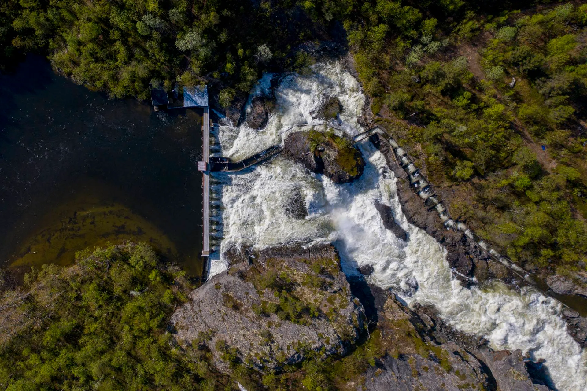 Luftfoto av en foss omgitt av skog og steiner med en gangbro over vannet.