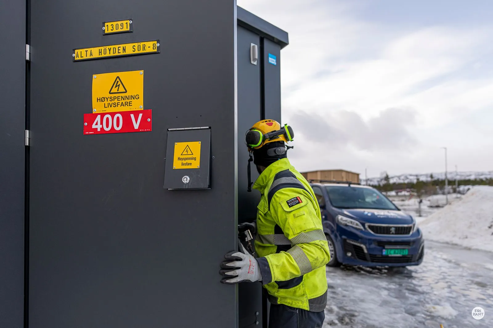En arbeider i gul sikkerhetsjakke og hjelm åpner døren til en elektrisk skap med advarsler om høy spenning 400 V, med en blå bil og snødekt bakgrunn.