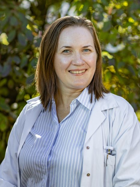 Dr. Natalie Gibis hat ein gestreiftes Shirt und einen weißen Laborkittel an und steht draußen mit grünem Laub im Hintergrund.