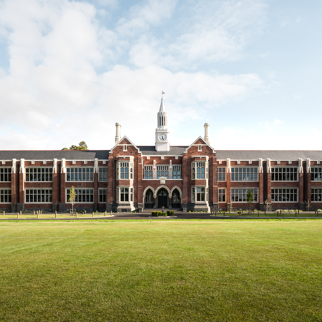 Front view of a large red brick school building with tall windows, clock tower, and green lawn.