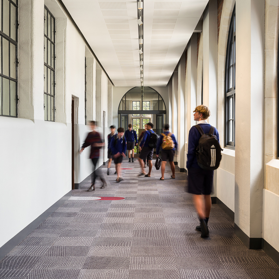 Students in school uniforms walk through a bright hallway with arched windows and patterned carpet.