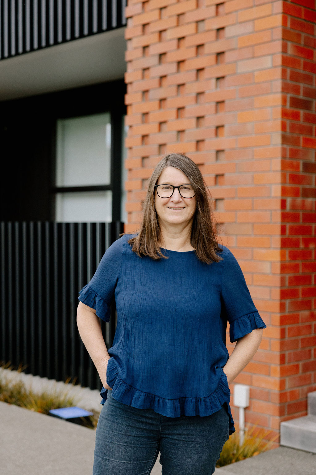 Woman with glasses wearing a blue blouse and jeans stands smiling in front of a brick wall.