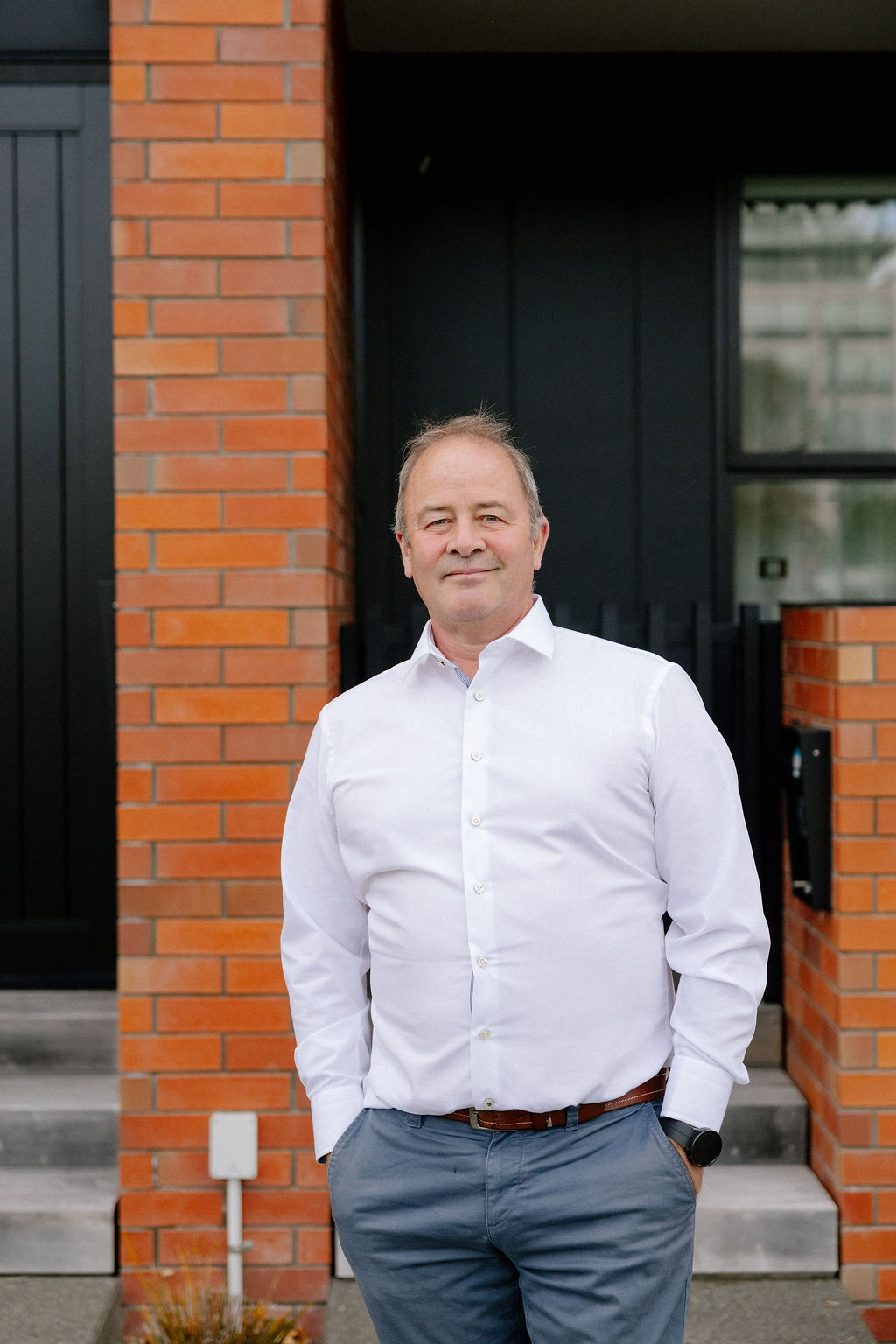 Middle-aged man wearing white shirt and blue pants stands with hands in pockets by brick house entrance.