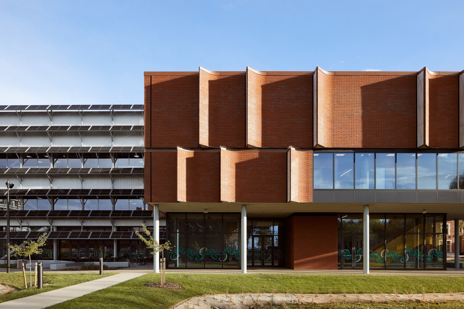 Modern brick and glass building with wavy facade sections and solar panels on adjacent structure.