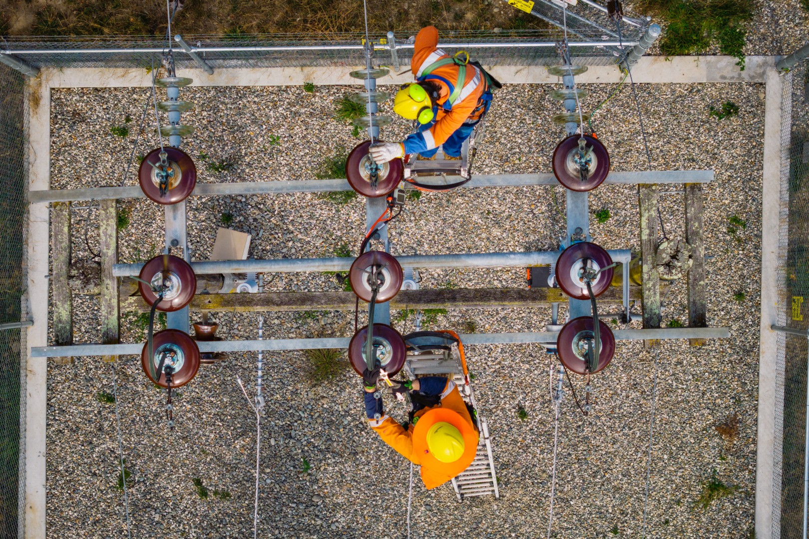 Two workers in orange safety gear repairing electrical insulators at a substation from above.