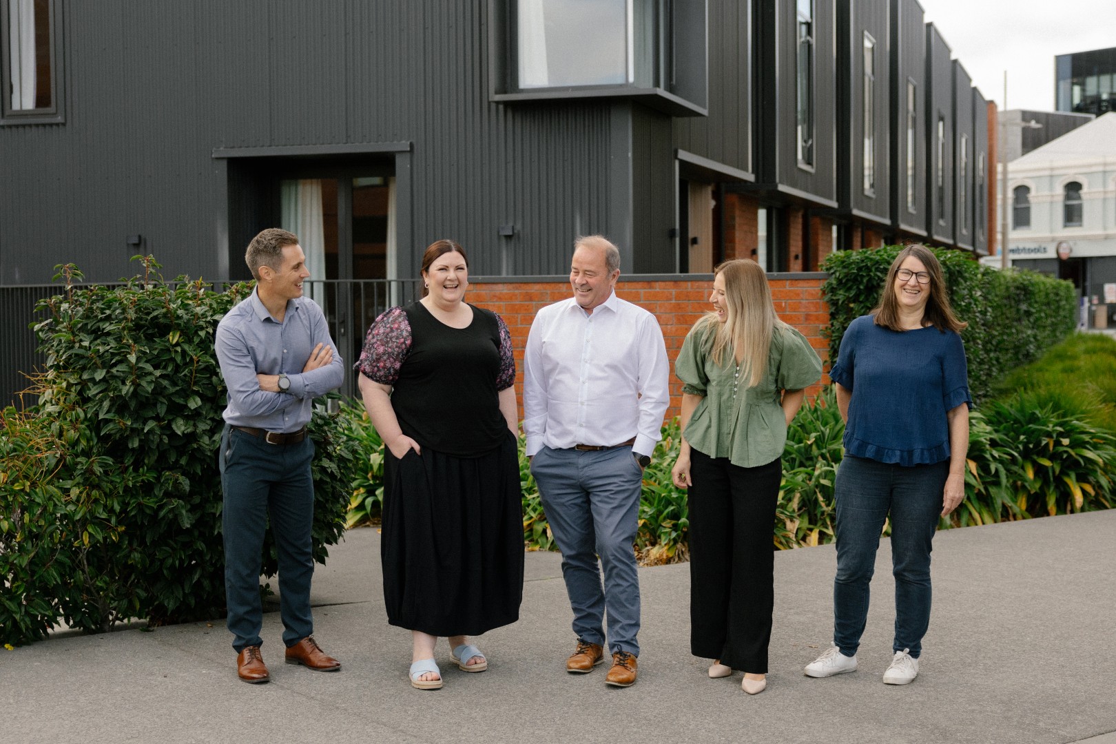 Five people standing outdoors near modern buildings, laughing and casually dressed.
