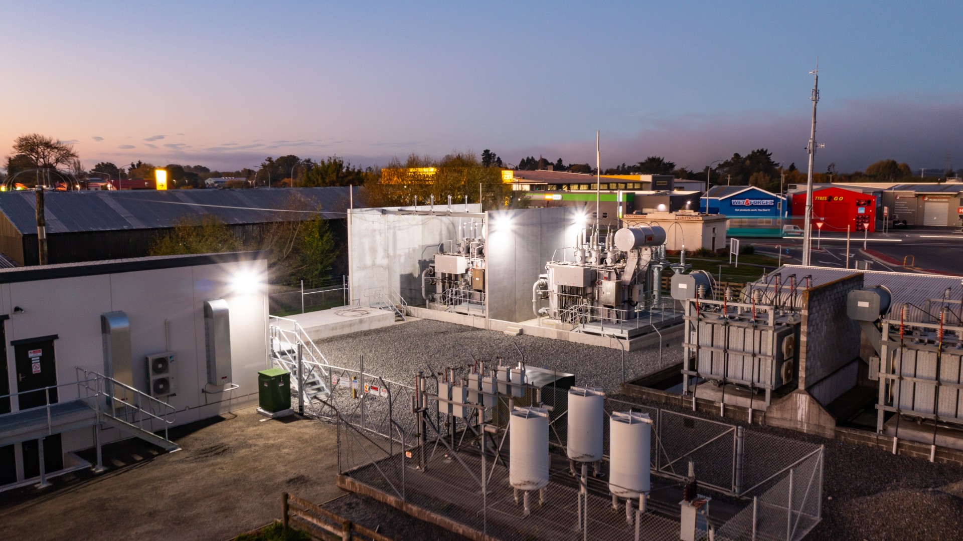 Industrial power substation with transformers and equipment fenced near commercial buildings at dusk.