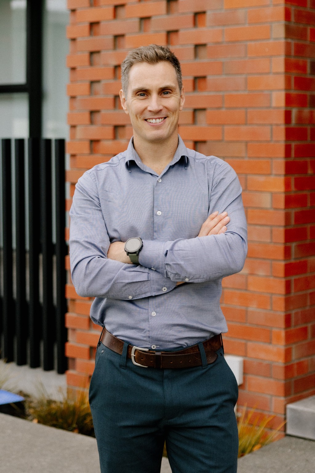 Man smiling with arms crossed wearing a light blue shirt and dark pants standing by a brick wall.