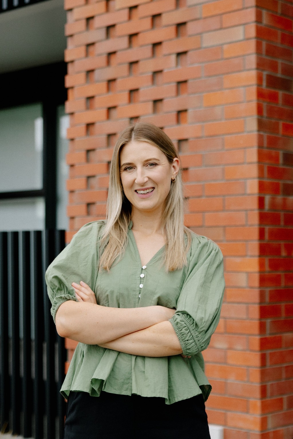 Smiling woman with blonde hair wearing a green blouse and standing with arms crossed outside.