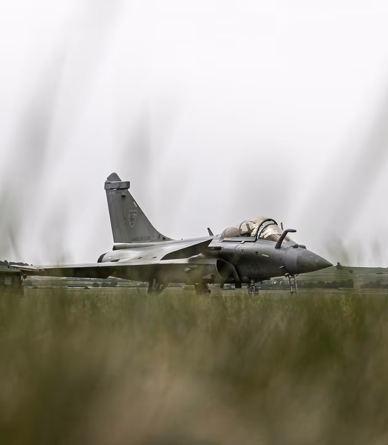 Avion de chasse militaire gris immobile sur une piste avec un ciel nuageux en arrière-plan et de l'herbe floue au premier plan.