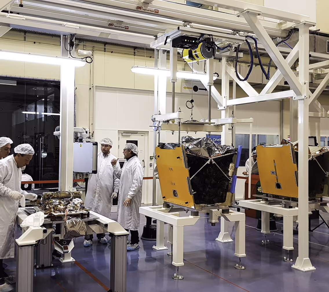 Techniciens en blouses blanches et filets à cheveux travaillent près d'équipements électroniques et de structures métalliques dans un laboratoire propre.