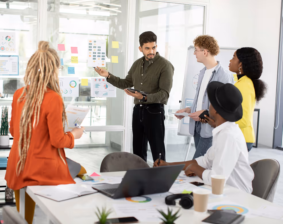 Un groupe de jeunes professionnels en réunion, discutant devant un tableau blanc et des graphiques collés sur une vitre.