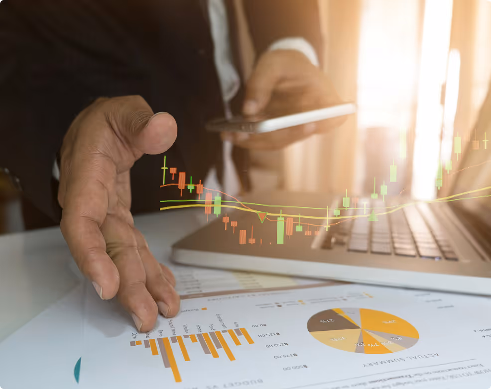 Une personne en costume consulte un smartphone devant un ordinateur portable avec des graphiques financiers et un diagramme circulaire imprimés sur une table.