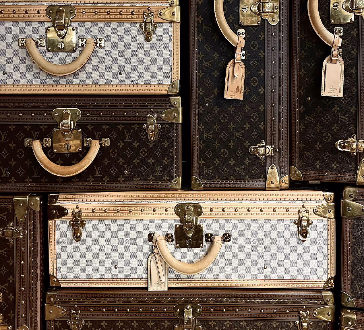 Gros plan sur une pile de malles Louis Vuitton avec motifs damiers et monogramme, poignées en cuir clair et quincaillerie dorée.