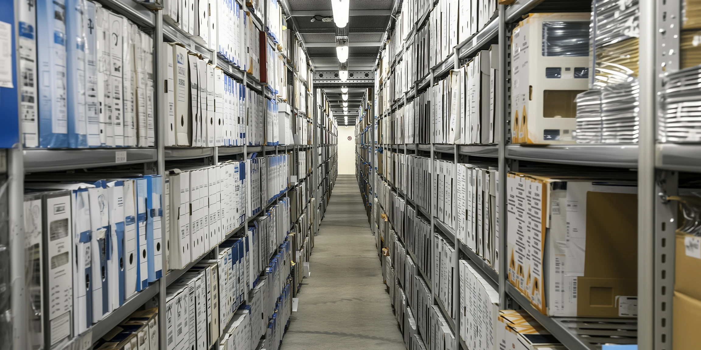 Long couloir d’archives avec des étagères métalliques remplies de dossiers et boîtes classées.