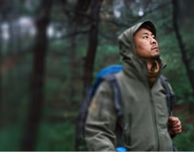 Man wearing a hooded jacket and carrying a blue backpack looking up in a forest.