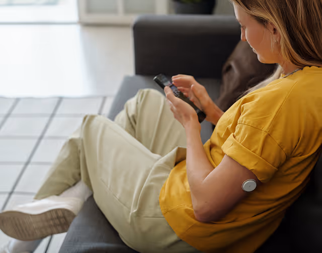 Woman sitting on a couch using a smartphone with a small medical device attached to her upper arm.