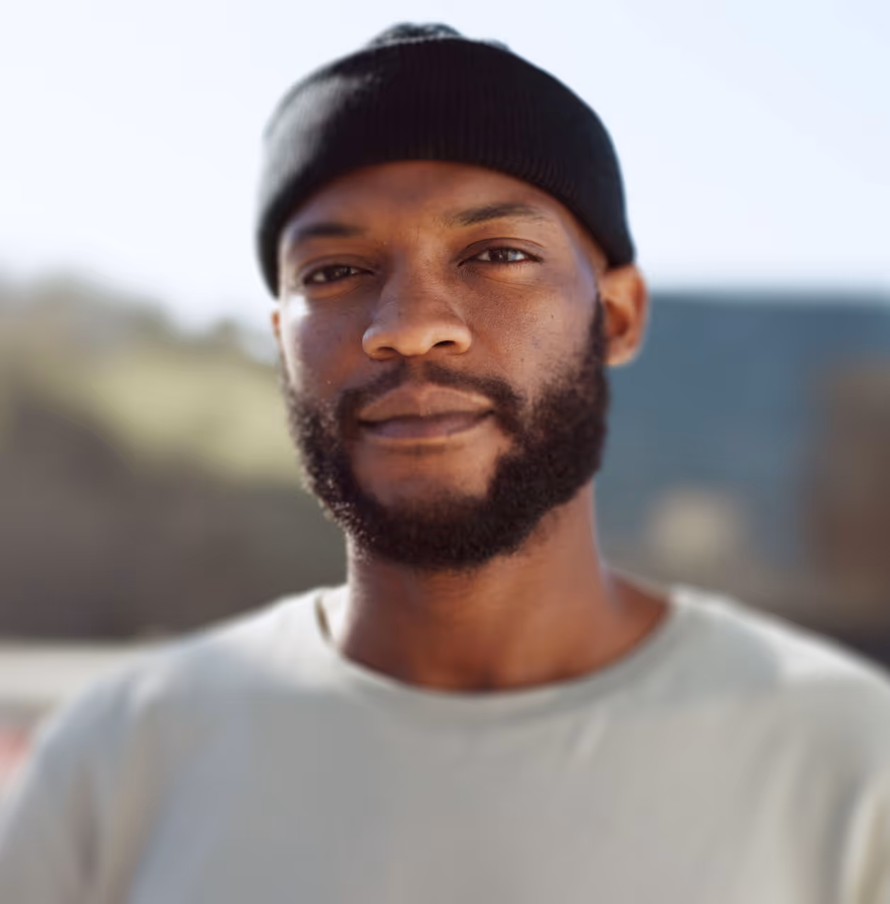 Close-up of a man with a beard wearing a black beanie and light-colored shirt outdoors.