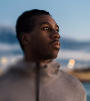 Close-up portrait of a man with a thoughtful expression looking to the side against a blurred outdoor background at dusk.