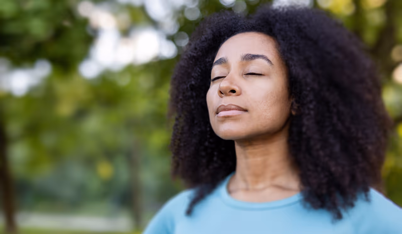 Woman with curly hair and closed eyes standing outdoors in a serene, green environment.