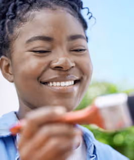 Smiling young woman holding a paintbrush outdoors with greenery in the background.