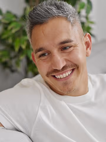 Smiling man with short gray hair and a mustache wearing a white t-shirt sitting on a couch.