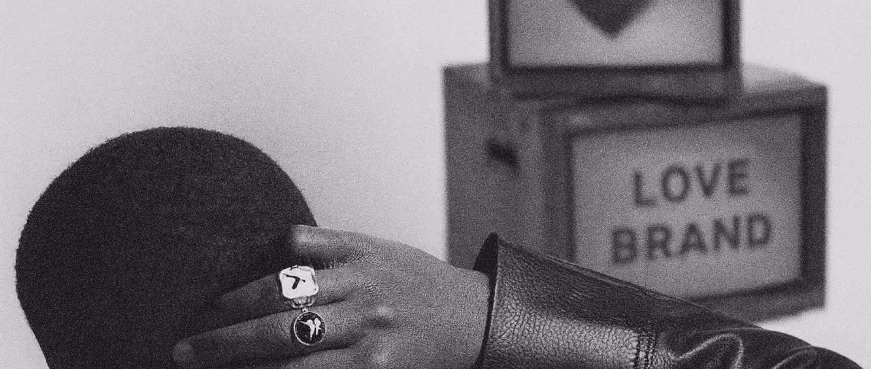 Une main portant deux bagues repose sur la tête d'une personne aux cheveux courts, avec des cubes en arrière-plan où est écrit "LOVE BRAND".