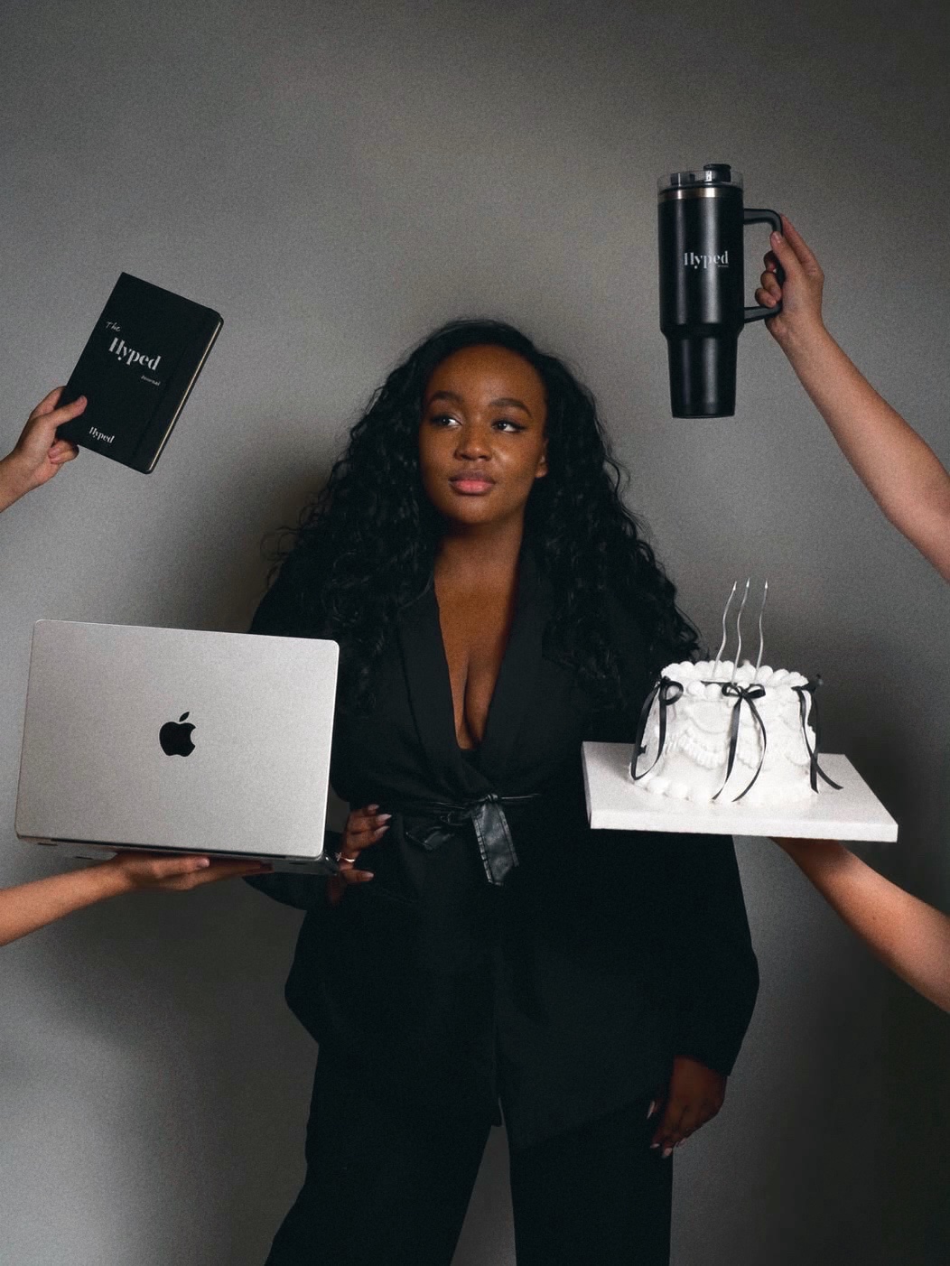 Fondatrice en costume noir entouree de mains lui tendant le carnet Hyped, une gourde Hyped, un MacBook et un gateau