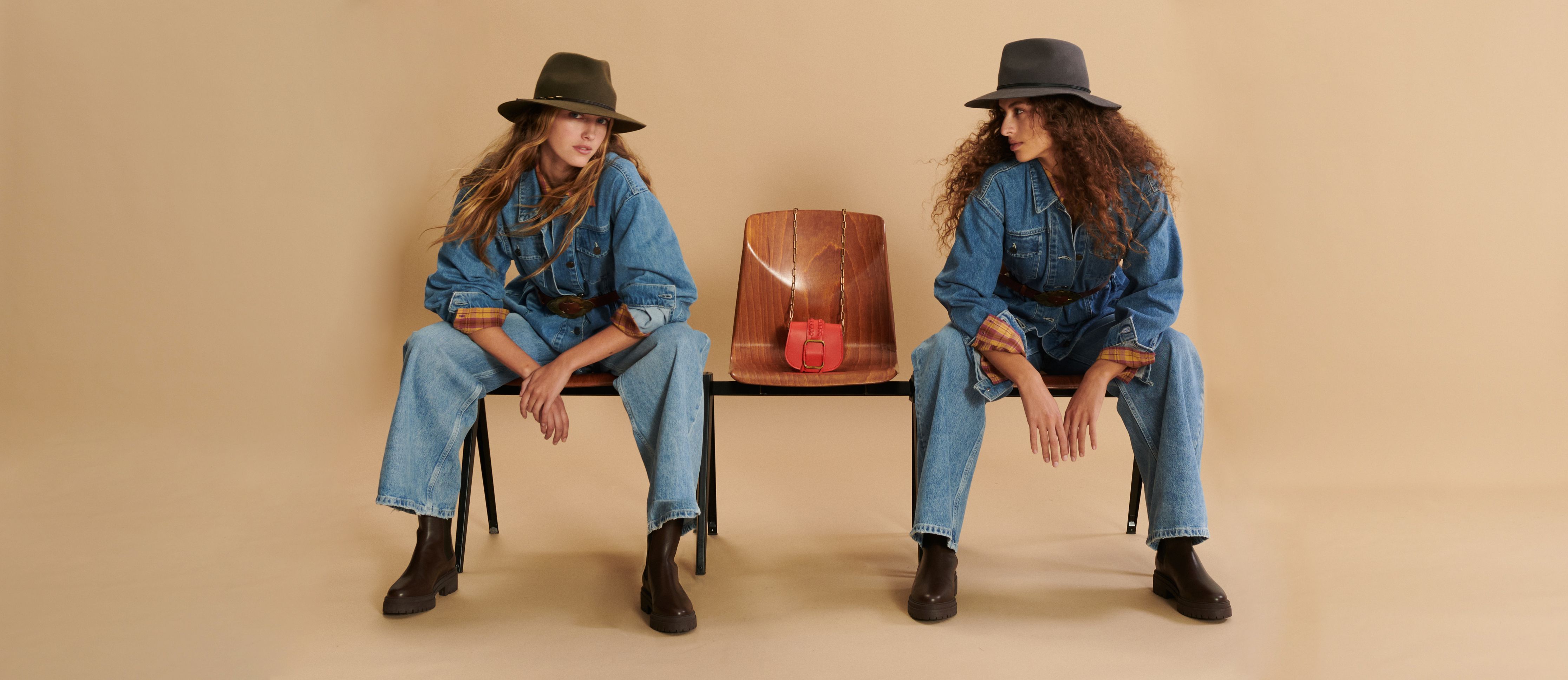 Two women sit on a bench wearing denim jumpsuits and brimmed hats
