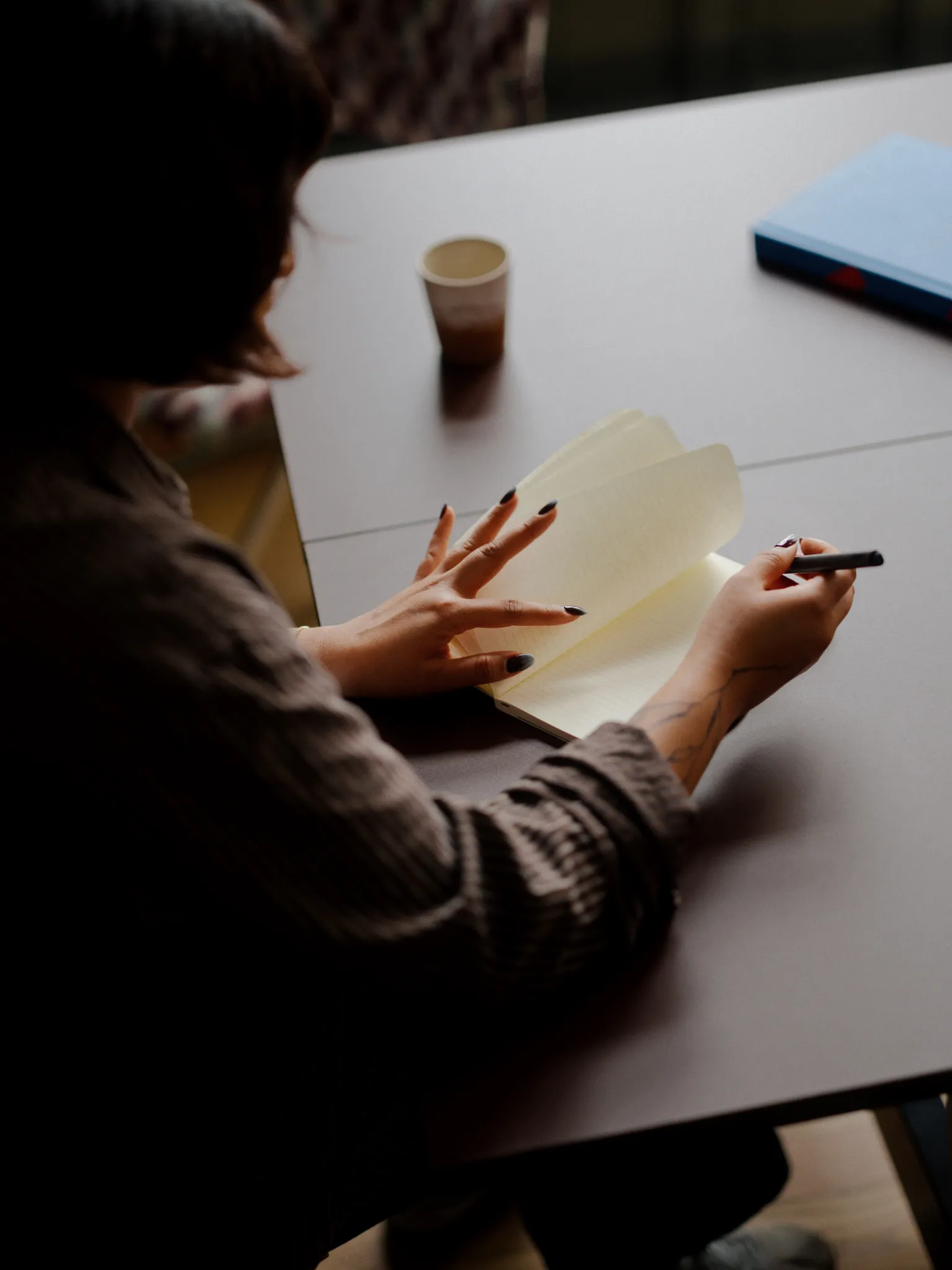 Person flipping through a blank notebook and holding a pen at a table with a cup and a closed blue book.